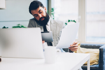 Busy bearded executive director solving working problems during mobile conversation via modern telephone while holding digital touch pad device and paper documentarion in hands sitting in office