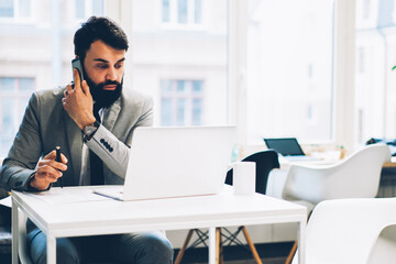 Concentrated bearded manager talking with client on smartphone while checking some information in online database on modern netbook sitting in office near publisity area for financial information