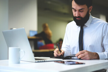 Concentrated bearded businessman planning working schedule noting points and details in notebook, prosperous manager using laptop computer for making researchers making accounting for financial report