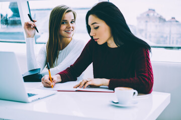 Portrait of attractive student with blonde hair smiling and looking at camera while another female writing article in notepad during preparation for upcoming exams sitting in university interior