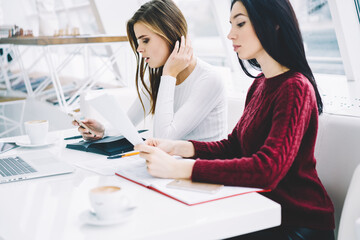 Two charming colleagues reading information and working together in office interior.Brunette and blonde female preparing for upcoming exams sitting with books and telephone in coworking space