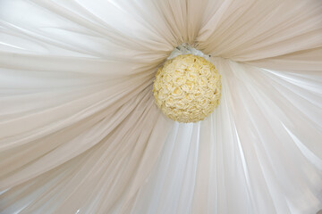 There is a white curtain on the ceiling . White rose ball on the ceiling .