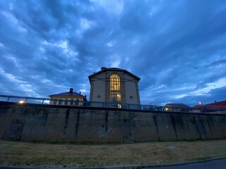 Gefängnisgebäude im Abendlicht. Großs Fenster ist gelb beläuchtet. Im Vordergrund Mauer mit Stacheldraht.