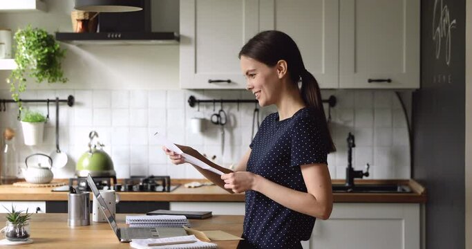Student girl standing in home kitchen takes out paper letter from envelope reading message feels excited by incredible great news. Job promotion or got reward, tax refund, bank loan acceptance concept