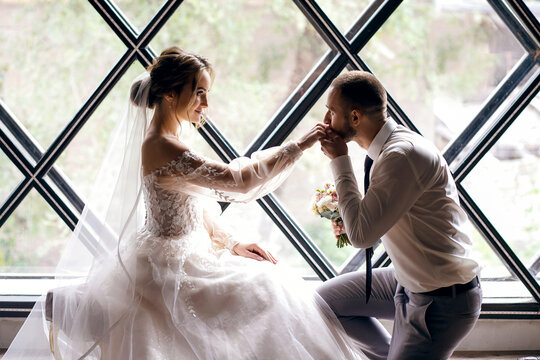 On the wedding day, a handsome groom on his knees kisses his wife hand against the background of a designer window, a man passionately looks into his woman eyes, wedding ceremony, horizontal photo