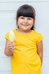 Adorable little girl with black hair eating ice cream, wearing a yellow shirt leaning against white...