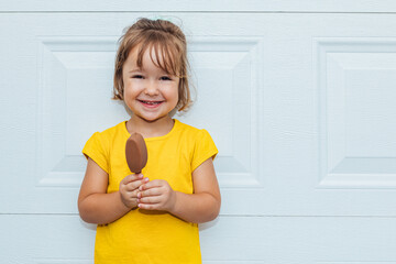 Adorable blond-haired girl eating ice cream, wearing a yellow shirt leaning against white background