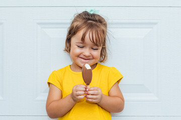 Adorable blond-haired girl eating ice cream, wearing a yellow shirt leaning against white background