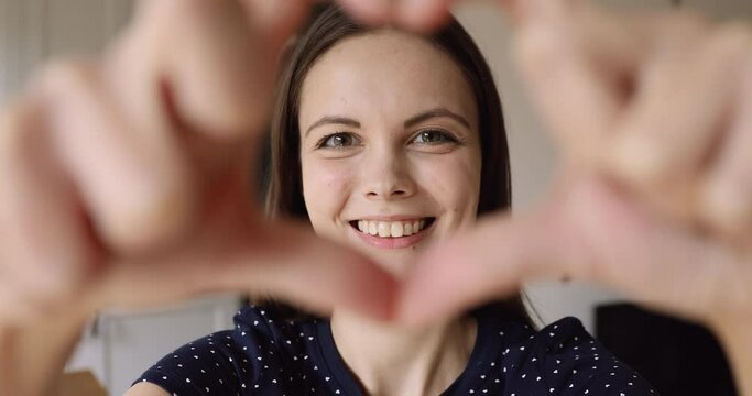 Close up happy girl makes with fingers heart shape smile look at camera through symbol. Satisfied client by services positive feedback, she in love romantic feelings, health insurance donation concept