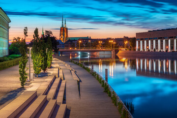 Evening on the boulevards on the Oder river in Wrocław. Illuminated historic buildings and bridges. Beautiful sky, light reflections on blurred water. Two cathedral towers in the distance