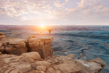Man stands triumphant with his hands outstretched in victory while standing on a cliff looking into the sunset and sunrise