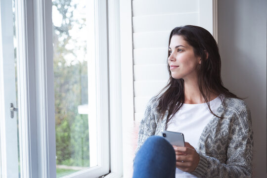 Woman with smartphone looking out of window at home