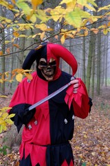 Obraz premium Halloween holiday concept. Evil harlequin in the autumn forest in the fog.A man in a suit of evil terrible jester with a knife. Evil clown cosplay costume.Autumn holidays. 