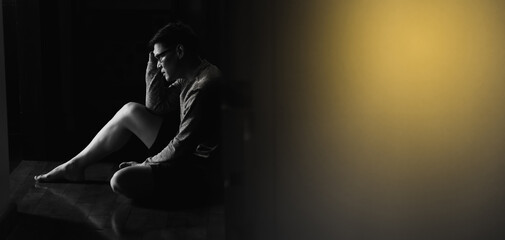 Desperate Asian young man sitting against a wall and looking down at home. Male on black background low key