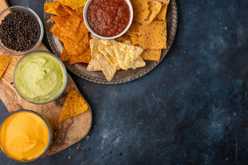 A plate of various snacks, potato chips, pita chips served with cheese, avocado, rosemary, and salt dip on a blue background. View from above. Copy space