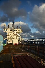 The deck of a large cargo ship with a tall white wheelhouse against a dark sky. The ship is illuminated by the bright evening sun. © German