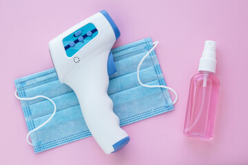 Non-contact infrared digital thermometer, hand sanitizer and medical mask on a pink background. A...