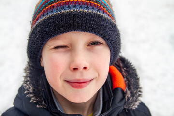 A teenage European boy in a hat and a warm jacket in winter. Close-up portrait. The child winks.