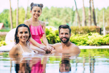 Handsome smiling man and cute excited woman with them little lovely daughter are having fun in summer swim pool while resting in hotel.