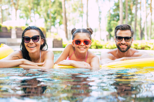 Handsome smiling man and cute excited woman with them little lovely daughter are having fun in summer swim pool while resting in hotel.