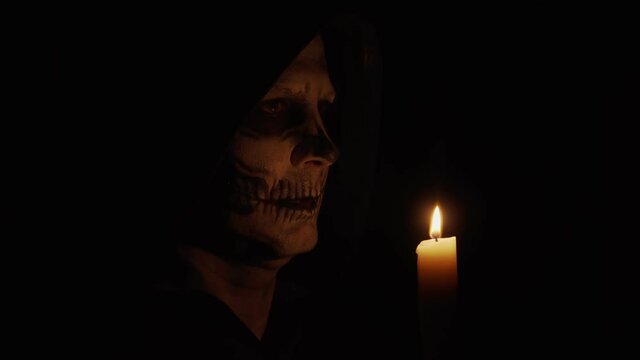 A man with skeletal makeup looks at the camera. The man is angry and casts a spell. Skeleton in the dim light of a candle. Halloween and horror concept.