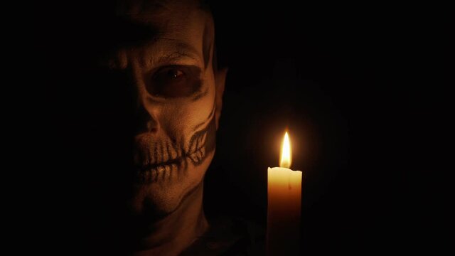 A man with skeleton makeup looks at the camera. A man smiles a creepy and funny smile. Skeleton in the dim light of a candle. Halloween and horror concept.