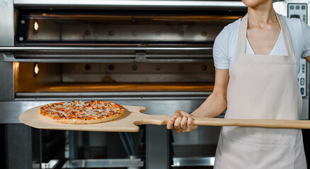 Young caucasian woman baker is holding a wood peel with fresh pizza near an oven at a baking manufacture factory.