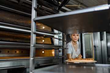 Young caucasian woman baker is holding a wood peel with fresh pizza near an oven at a baking manufacture factory and putting it on a metal shelves rack.