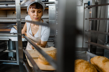 Close up of young caucasian woman baker putting the fresh bread on the shelves/rack at baking manufacture factory.