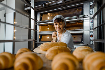 Close up of young caucasian woman baker putting the fresh bread on the shelves/rack at baking manufacture factory.