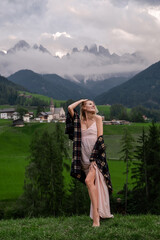 Naklejka premium Dolomites. Santa Magdalena. Italy. Smiling blonde woman in coffee dress & checkered shirt poses on background of alpine village, hills, green meadow & high mountains peak wrapped by the clouds