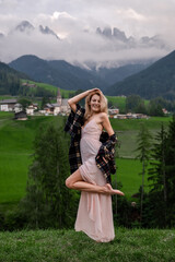 Dolomites. Santa Magdalena. Italy. Smiling blonde woman in coffee dress poses with raised leg & hand on background of alpine village, hills, green meadow & high mountains peak wrapped by the clouds