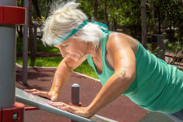 Slender elderly woman with gray hair does push-ups on a simulator in a city park during individual fitness classes. Close-up