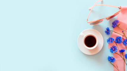 Top view of a cup of coffee with blue cornflowers, pink sunglasses and pink silk ribbon on a pastel blue background. Summer beach flat lay. Lifestyle, copy space, mockup.