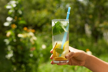 Faceless woman holding glass of cocktail with lemon and mint on green garden background, glass of cold summer beverage, refreshing in heat.