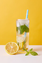 Lemonade with lemon, mint and ice cubes, juicy beverage in glass with straw, refreshing summer drink on table decorated with lemon piece.