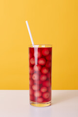 Glass cup of red cocktail with drinking tube and cherries isolated over yellow background, fresh non alcoholic summer beverage on table.