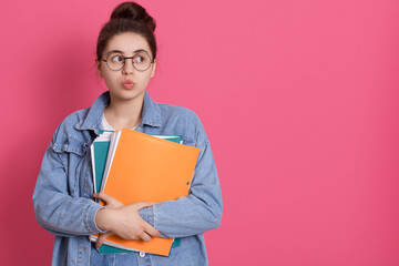 Thoughtful pretty woman looks pensively aside with rounded lips, holds paper folder, has pleasant appearance, being deep in thoughts. Copy space for advertisement.