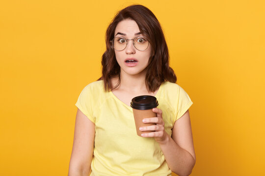 Astonished displeased woman with dark hair, holding paper cup, wearing yellow t shirt, holds disposable cup of coffee, being upset and shocked, posing with opened mouth and big eyes.