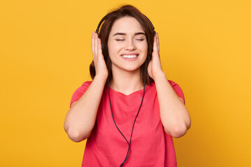 Young smiling brunette girl with closed eyes wearing red t-shirt, listening music via headphones, enjoying favorite songs and relaxing, isolated over yellow background.