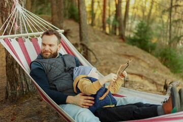 Young family hipster playing with son on a hammock in park on camping trip, family vacation