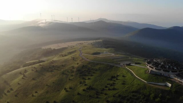 Aerial view of the Buzludzha monument in Bulgaria at sunrise, abandoned communist concrete structure with a red star on a green mountain hill with wind turbines in the background
