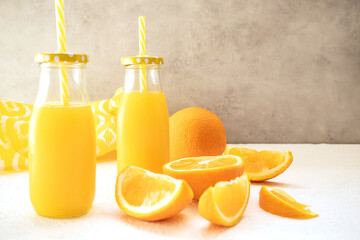 Two glass bottles with caps and cocktail tubes with fresh orange juice stand on a white table against a gray concrete wall. Juicy slices of orange lie near. The concept of a healthy diet, detox diet.