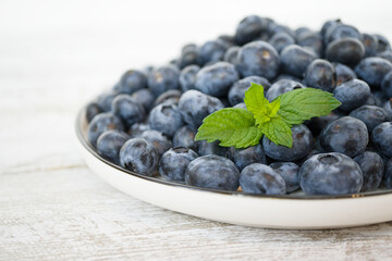 A full white bowl of juicy and tasty blueberries stands on a light wooden table. A bowl is cropped with fresh mint leaves. Seasonal healthy berries, proper nutrition. Close-up