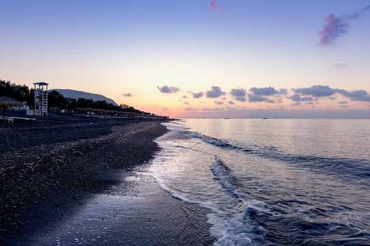 Santorini, black pebble beach of Perivolos, Perisa at sunrise. Sea waves, beach chairs and umbrellas, beautiful sky with clouds.