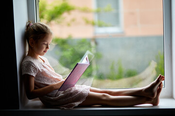 Cute little girl reading book at home, at windowsill