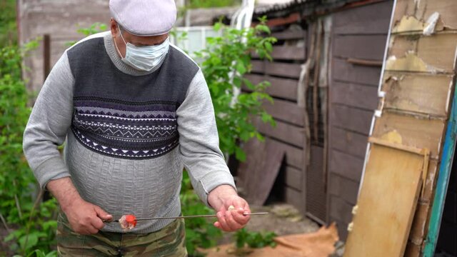 Mature man in medical mask impales raw shashlik on skewer. Older male holds skewer with barbecue. Concept of picnic outdoor.