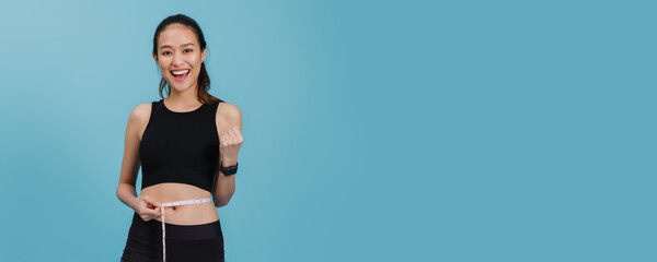 Portrait of confident beautiful asian fitness woman standing and use tape measure to measure waist  after exercise with smiling face on blue colour background.Concept of slim and healthy happy girl.