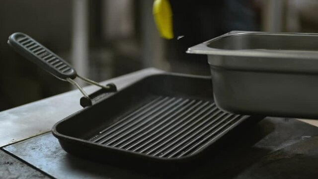 Young cook smears pan with oil on stove while working in restaurant kitchen irrl. Male chef applies butter to hot metal surface before cooking while standing indoors. Professional is in work process