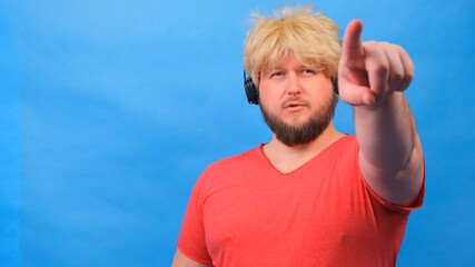 Naklejka premium freaky fat man in a wig and a pink T-shirt in headphones shows a finger on the camera at you against a blue background.